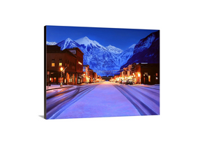 A canvas print depicting a snowy Main Street in Telluride, Colorado, with illuminated lights and snow-covered mountains in the background. Cityscape art by wildlife artist James Corwin.