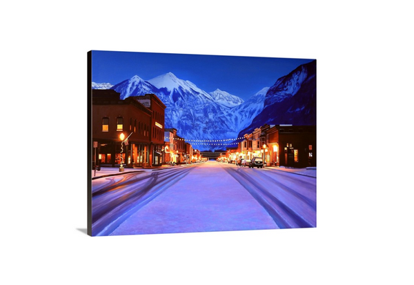 A canvas print depicting a snowy Main Street in Telluride, Colorado, with illuminated lights and snow-covered mountains in the background. Cityscape art by wildlife artist James Corwin.